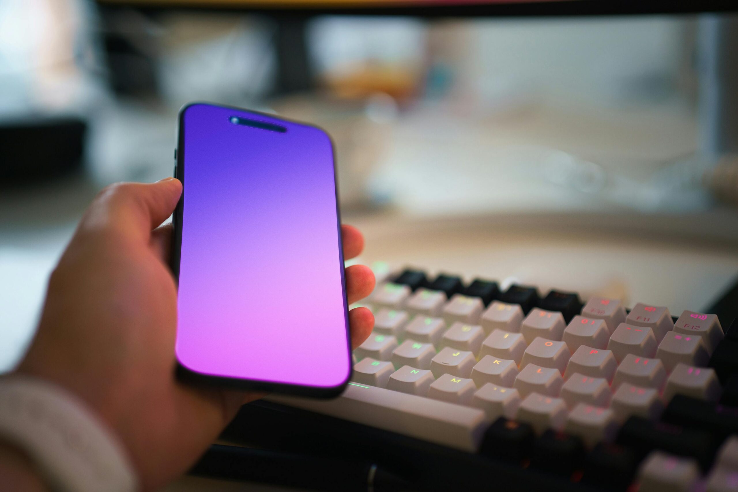 Hand holding a smartphone with a blank purple screen, positioned near a colorful keyboard on a desk.